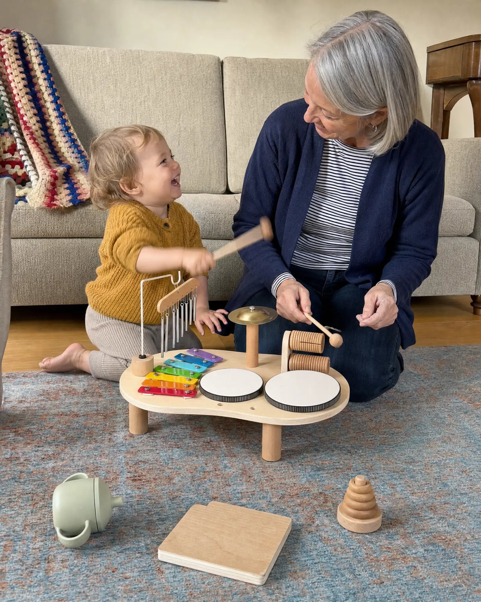 A grandmother and a toddler on the living room floor, each holding a wooden mallet at the music table