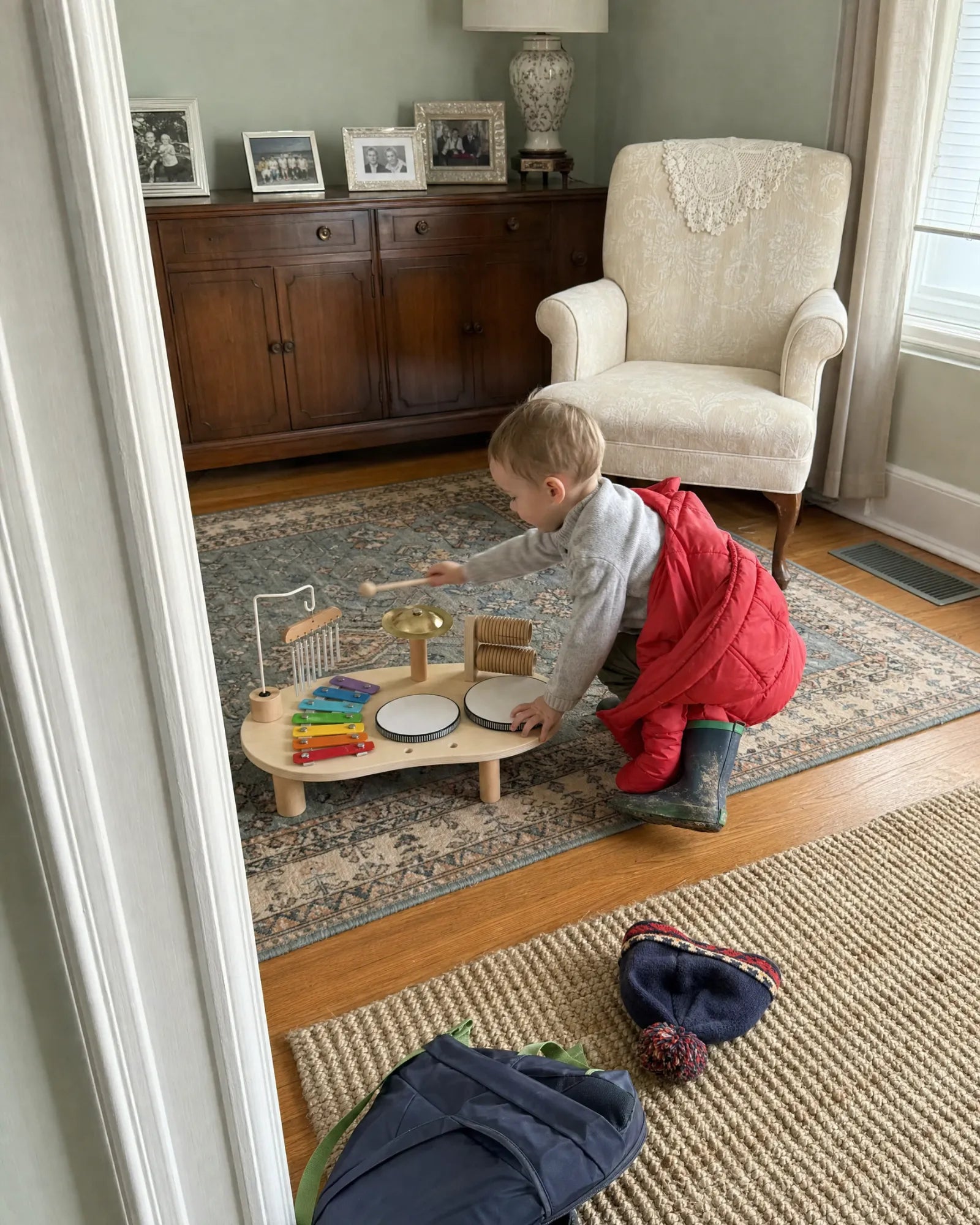 A toddler with a red coat half off reaching a mallet toward the cymbal on the music table