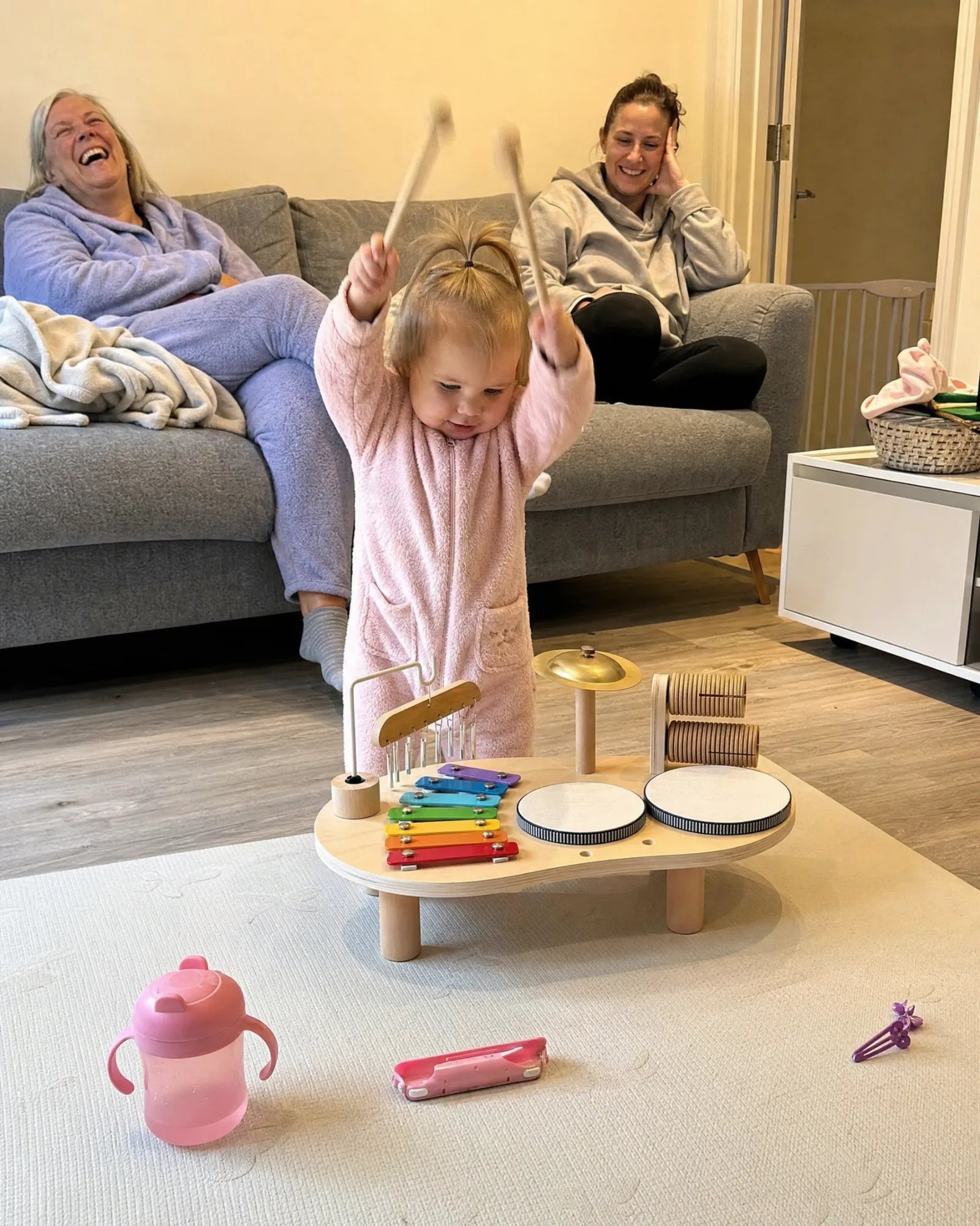 A toddler with both mallets raised over the drums while two women laugh on the sofa behind